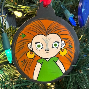 May include: Hand-painted Christmas ornament featuring a cartoon character with orange hair, green eyes, and a green shirt. The ornament is round and has a black background.