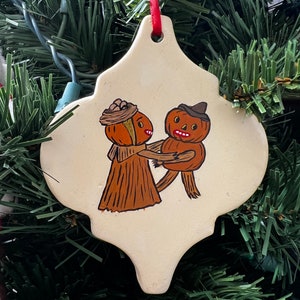 Peut inclure: Une décoration de Noël en céramique blanche en forme de losange avec une image peinte de deux personnages en forme de citrouille se tenant la main. La femme citrouille porte une robe brune et un chapeau de paille avec un ruban brun. L'homme citrouille porte un chapeau brun et un gilet brun.