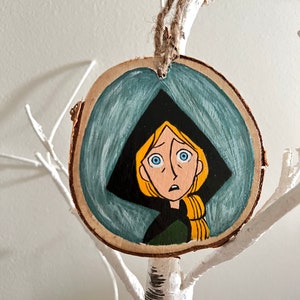 May include: A hand-painted wooden ornament featuring a cartoon character with blonde hair, blue eyes, and a worried expression. The character is wearing a black hooded cloak and a green shirt. The ornament is painted on a round piece of wood with a rustic finish.