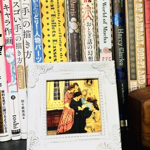 Puede incluir: Una imagen enmarcada en blanco con una ilustraci&oacute;n vintage de cuatro mujeres en una habitaci&oacute;n. Las mujeres llevan vestidos largos y est&aacute;n reunidas alrededor de una mesa. La imagen est&aacute; colocada en un estante con una fila de libros detr&aacute;s.
