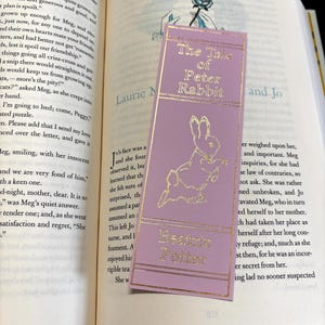 May include: A purple bookmark with gold lettering featuring the title "The Tale of Peter Rabbit" by Beatrix Potter. The bookmark features a rabbit illustration.