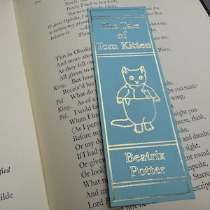 May include: A blue bookmark with gold lettering featuring a cartoon illustration of a kitten. The bookmark reads "The Tale of Tom Kitten" and "Beatrix Potter".