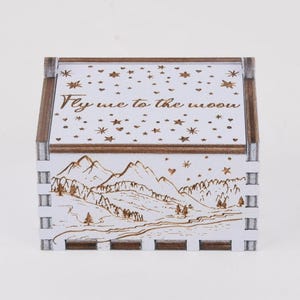 Może przedstawiać: Białe drewniane pudełko z motywem gór i gwiazd. Wieko zdobi napis "Fly me to the moon" oraz wycięcia w kształcie gwiazd. Pudełko ma brązową drewnianą ramę i jest przedmiotem dekoracyjnym.