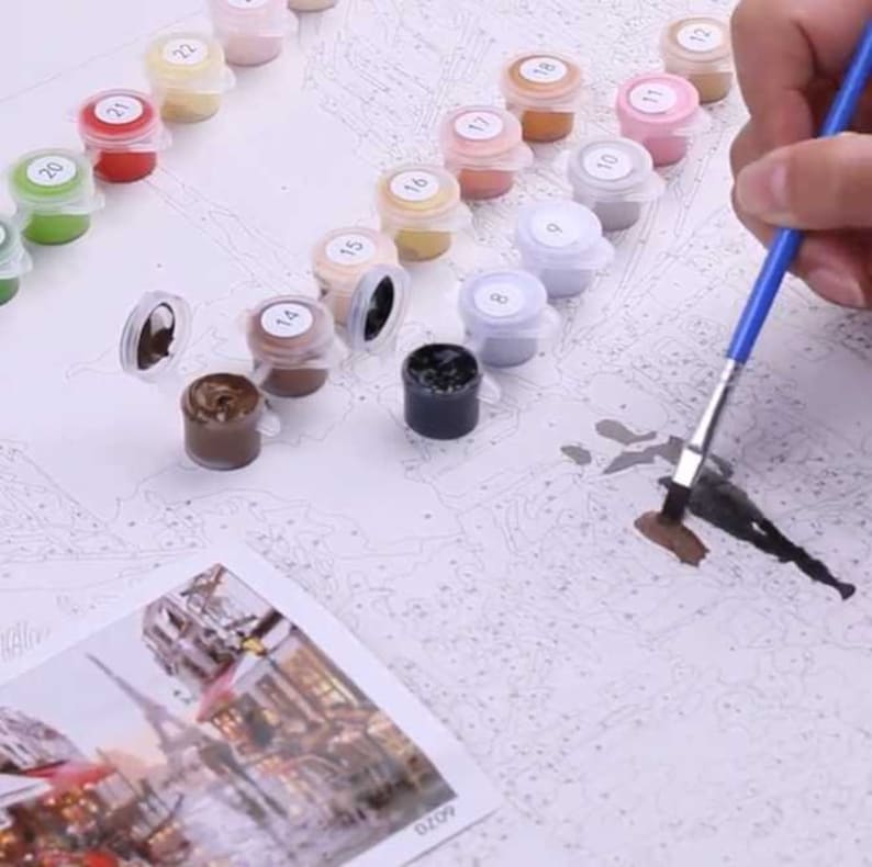 May include: A paint by numbers kit with a variety of paint colors in small containers. The containers are labeled with numbers from 1 to 22. A person is using a paintbrush to apply paint to the canvas.