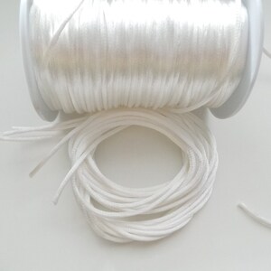 May include: A spool of white cord, partially unraveled, sits on a white surface. The cord is tightly wound on the spool and also coiled in a separate pile. The cord appears to be made of a smooth, synthetic material.