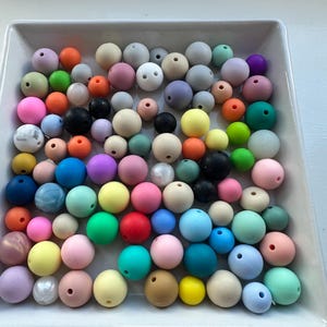 Peut inclure: Un ensemble de perles en silicone colorées dans un plat carré blanc. Les perles sont de différentes nuances de bleu, vert, rose, jaune, orange et marron.