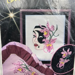 May include: A cross-stitch pattern kit featuring an orchid mask design. The kit includes a pillow with a pink and white orchid design, a round black and white pillow with a pink orchid design, and a framed cross-stitch pattern with a black and white mask with pink orchids. The kit is titled "Orchid Mask" and "Jeanne Christine MM 0009".