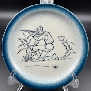 May include: A blue and white ceramic plate with a black and white illustration of a hunter with a gun and a dog. The hunter is kneeling and aiming the gun. The dog is standing next to him.