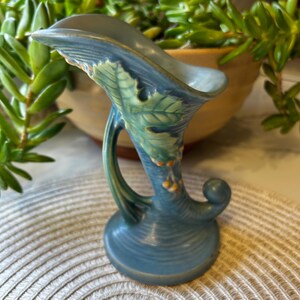 May include: A blue ceramic cornucopia vase featuring a raised green leaf design and small orange berry accents. The vessel has a wide, flared opening, a curved body, and a circular base, with a textured surface throughout the exterior.