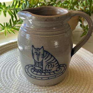 May include: A stoneware pitcher with a light gray speckled finish. A blue cat design is painted on the front. The pitcher has a curved handle and a spout for pouring. The cat is sitting on a plate that says "milk".