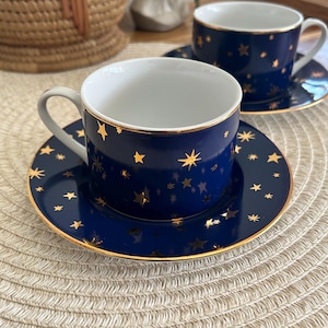 May include: Two dark blue ceramic teacups and matching saucers featuring a gold star pattern. Each cup has a white interior and a gold-trimmed rim. The saucers also feature a gold-trimmed edge and a scattered gold star design across the surface.