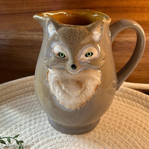 May include: A ceramic pitcher with a fox face design. The pitcher is primarily gray with a white and tan chest and green eyes. The handle is curved and the interior is a golden brown color. The pitcher is sitting on a woven mat.