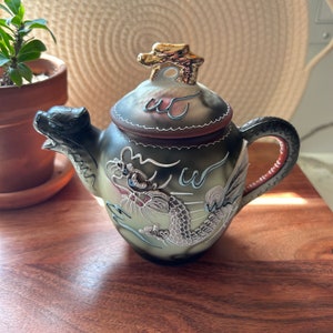 May include: A small, black and white ceramic teapot with a dragon design. The teapot has a gold lid with a dragon finial. The teapot is sitting on a wooden surface.