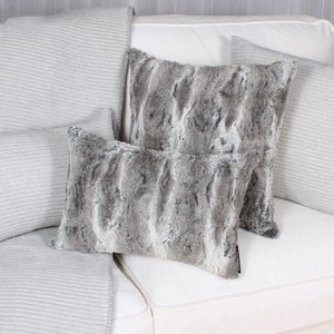May include: Three decorative throw pillows on a white sofa. The pillows are made of a soft, grey faux fur fabric. The sofa is covered in a white fabric with a subtle texture.