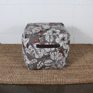May include: A gray and white patterned square ottoman with a black leather handle. The ottoman has a tropical print with birds and flowers.