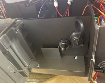 May include: Close-up of a black plastic box with a black toggle switch and a black connector with a black strap. The box is attached to a larger black plastic object.