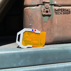 Puede incluir: Un dispositivo de pl&aacute;stico blanco y naranja con una pantalla que muestra el texto "74E1V", "DATA FILE TRANSFER" y "SUM FORCE LEGION". El dispositivo est&aacute; sobre una superficie verde.