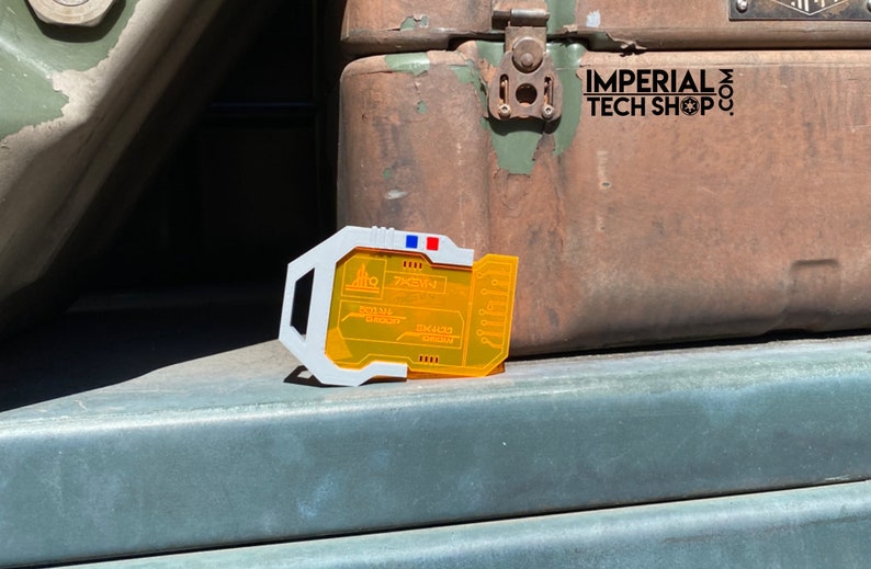 Puede incluir: Un dispositivo de pl&aacute;stico blanco y naranja con una pantalla que muestra el texto "74E1V", "DATA FILE TRANSFER" y "SUM FORCE LEGION". El dispositivo est&aacute; sobre una superficie verde.