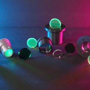 May include: An assortment of optical components, including lenses and metal housings, are arranged on a dark surface. The lenses vary in color, with some appearing green and others clear or black. The scene is illuminated with blue and pink light.