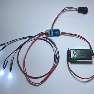 K&ouml;nnte beinhalten: Eine Platine mit Dr&auml;hten, die mit vier wei&szlig;en LED-Leuchten, einem Batteriepack und einem Druckknopfschalter verbunden sind.
