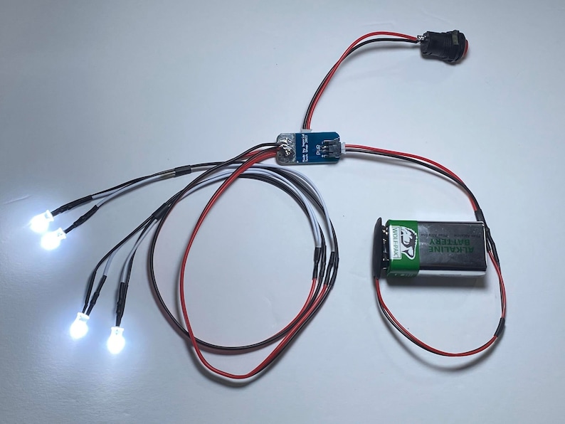 K&ouml;nnte beinhalten: Eine Platine mit Dr&auml;hten, die mit vier wei&szlig;en LED-Leuchten, einem Batteriepack und einem Druckknopfschalter verbunden sind.