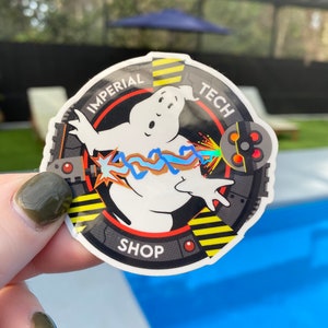 Può includere: Un adesivo circolare con un fantasma dei cartoni animati che emette un raggio di energia blu e marrone. L'adesivo presenta il testo "IMPERIAL TECH SHOP" in un design nero e giallo, con un bordo grigio e rosso.