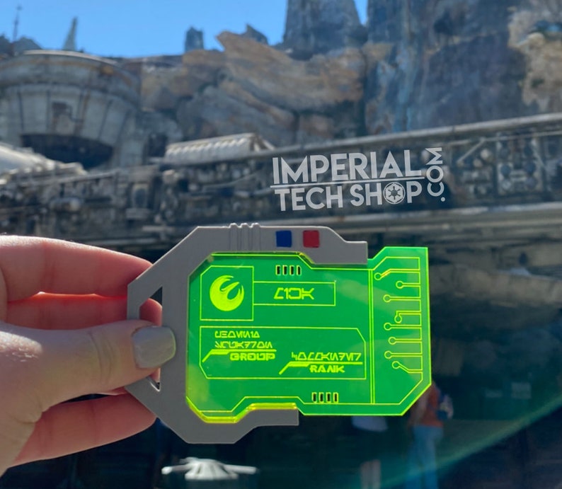 Puede incluir: Una tarjeta de pl&aacute;stico verde y gris con una pantalla verde brillante. La pantalla muestra el texto "L03W1A GROUP" y "H0C0K1A77 RANK". La tarjeta tambi&eacute;n tiene un peque&ntilde;o s&iacute;mbolo blanco que se asemeja a un ala de p&aacute;jaro. La tarjeta est&aacute; delante de un fondo borroso de un edificio con el texto "IMPERIAL TECH SHOP" en &eacute;l.