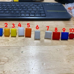 Pu&ograve; includere: Dieci piccoli bottoni quadrati di diversi colori, numerati da 1 a 10. I bottoni sono disposti in una fila da sinistra a destra. I colori sono blu, giallo, bianco, arancione, bianco, bianco, rosso, blu e rosso.