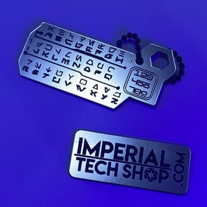 Pode incluir: Um chaveiro de metal prateado com uma corrente preta. O chaveiro tem uma pequena etiqueta de metal com o texto "IMPERIAL TECH SHOP.COM" e uma etiqueta maior com uma grade de símbolos e números. Os números na etiqueta são 129, 450 e 780.