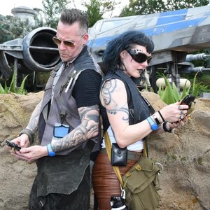 Puede incluir: Dos personas con tatuajes se ponen de espaldas frente a una gran nave espacial gris. La persona de la izquierda lleva un chaleco gris y una pulsera azul. La persona de la derecha lleva un chaleco negro y una pulsera azul. Ambas personas miran sus tel&eacute;fonos.