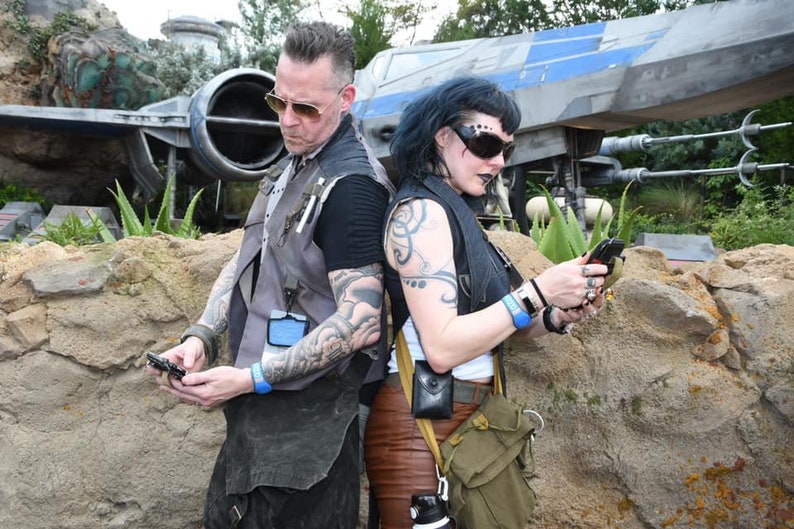 Puede incluir: Dos personas con tatuajes se ponen de espaldas frente a una gran nave espacial gris. La persona de la izquierda lleva un chaleco gris y una pulsera azul. La persona de la derecha lleva un chaleco negro y una pulsera azul. Ambas personas miran sus tel&eacute;fonos.