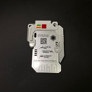 May include: A silver-colored, rectangular, plastic card with a black and white barcode and text that reads "17-14818 ID#  HEXA771NK ORIGIN  JTMOL VIAMKIA RANK". The card has a black and white symbol that resembles a circle with a triangle inside. The card has a red and green light on the top.