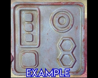 Könnte beinhalten: Eine graue Metallplatte mit rostiger Oberfläche. Die Platte hat vier runde Knöpfe, einen Kreis und zwei sechseckige Formen. Der Text "EXAMPLE" ist unten auf der Platte aufgedruckt.