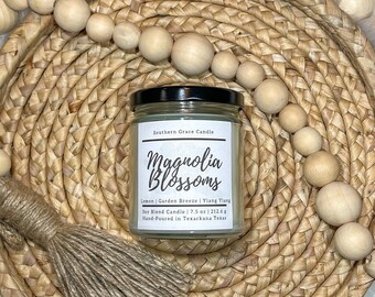 May include: A white candle jar with a black lid and a white label that reads "Southern Grace Candle Magnolia Blossoms Lemon | Garden Breeze | Ylang Ylang Soy Hand Candle | 7.5 oz | 212.6 g Hand-Poured in Texarkana, Texas" on a woven straw tray with wooden beads.