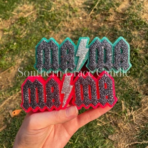 May include: Two glittery, decorative air fresheners in the shape of the word "MAMA" with a lightning bolt. One is neon pink with black glitter, the other is teal with black glitter. The text "Southern Grace Candle" is also visible.