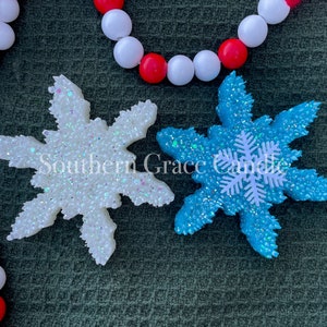 May include: Two sparkly snowflake-shaped air fresheners, one white and one blue, with a white snowflake design on the blue one. The text "Southern Grace Candle" is visible in the image.