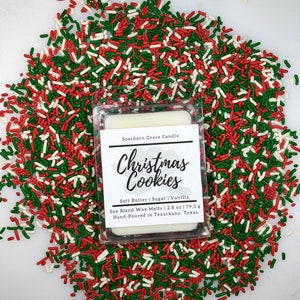 May include: A white wax melt with a clear plastic container. The label reads "Southern Grace Candle Christmas Cookies Soft Butter | Sugar | Vanilla Soy Blend Wax Melts | 2.8 oz | 79.3 g Hand-Poured in Texarkana, Texas". The wax melt is surrounded by red, green, and white sprinkles.