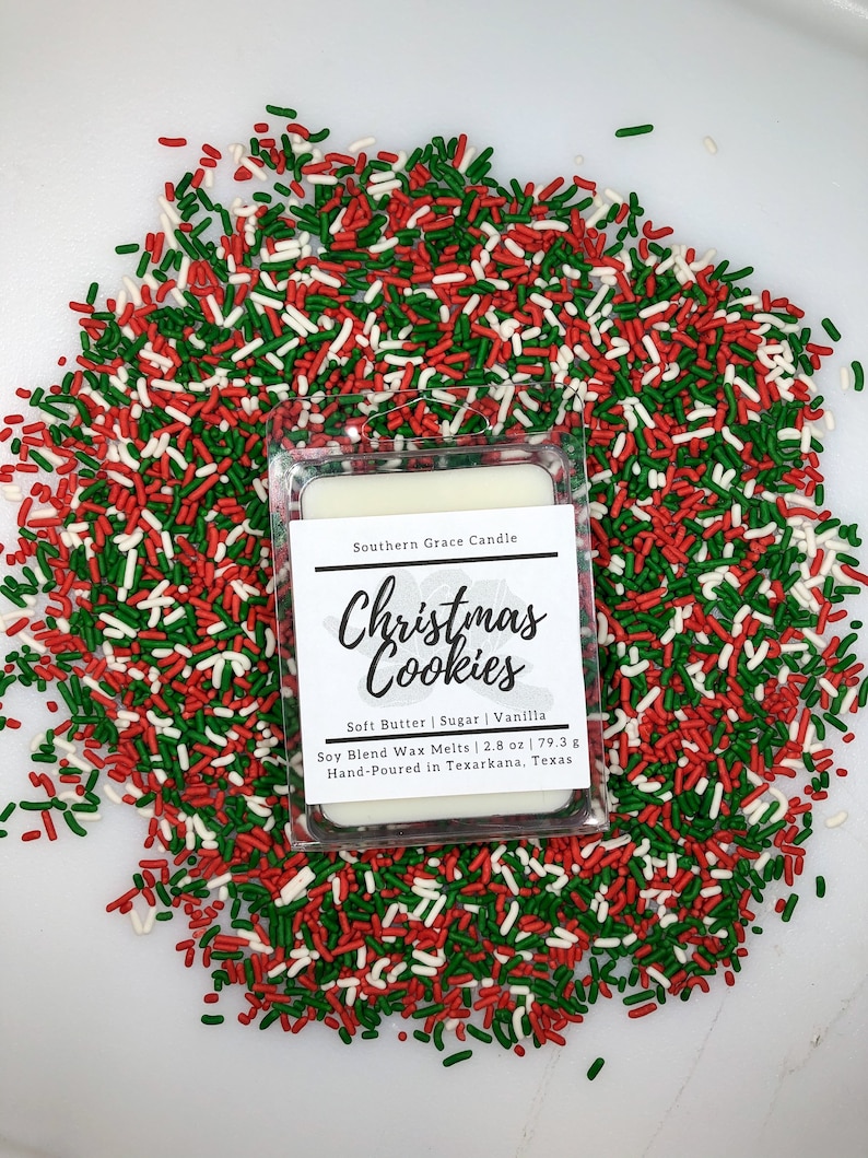 May include: A white wax melt with a clear plastic container. The label reads "Southern Grace Candle Christmas Cookies Soft Butter | Sugar | Vanilla Soy Blend Wax Melts | 2.8 oz | 79.3 g Hand-Poured in Texarkana, Texas". The wax melt is surrounded by red, green, and white sprinkles.