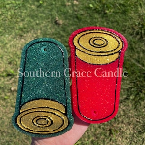 May include: Two handmade shotgun shell candles, one green and one red, with gold accents. The candles are shaped like shotgun shells and have a black outline. The text "Southern Grace Candle" is visible on the image.