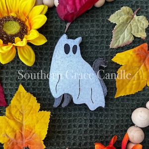 May include: A light blue ghost-shaped decoration with black eyes and a black tail, surrounded by fall-themed decorations. The image includes a yellow sunflower, orange and yellow leaves, and wooden beads. The text "Southern Grace Candle" is visible.