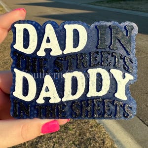 May include: A blue and white decorative sign with the text "DAD IN THE STREETS DADDY IN THE SHEETS". The sign has a textured appearance and is held by a person with pink nail polish.