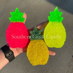May include: Three pineapple-shaped car air fresheners in pink, yellow, and green. The fresheners are made of a textured material and have a green pineapple top.