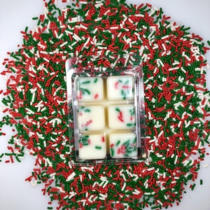May include: Six white wax melts in a clear plastic tray, surrounded by red, green, and white sprinkles. The wax melts are decorated with red and green sprinkles.