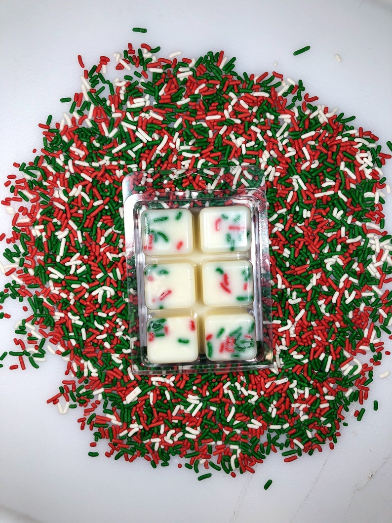 May include: Six white wax melts in a clear plastic tray, surrounded by red, green, and white sprinkles. The wax melts are decorated with red and green sprinkles.