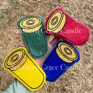 May include: Four shotgun shell candle holders in different colors: green, red, blue, and yellow. Each holder has a gold-colored top with a black outline.