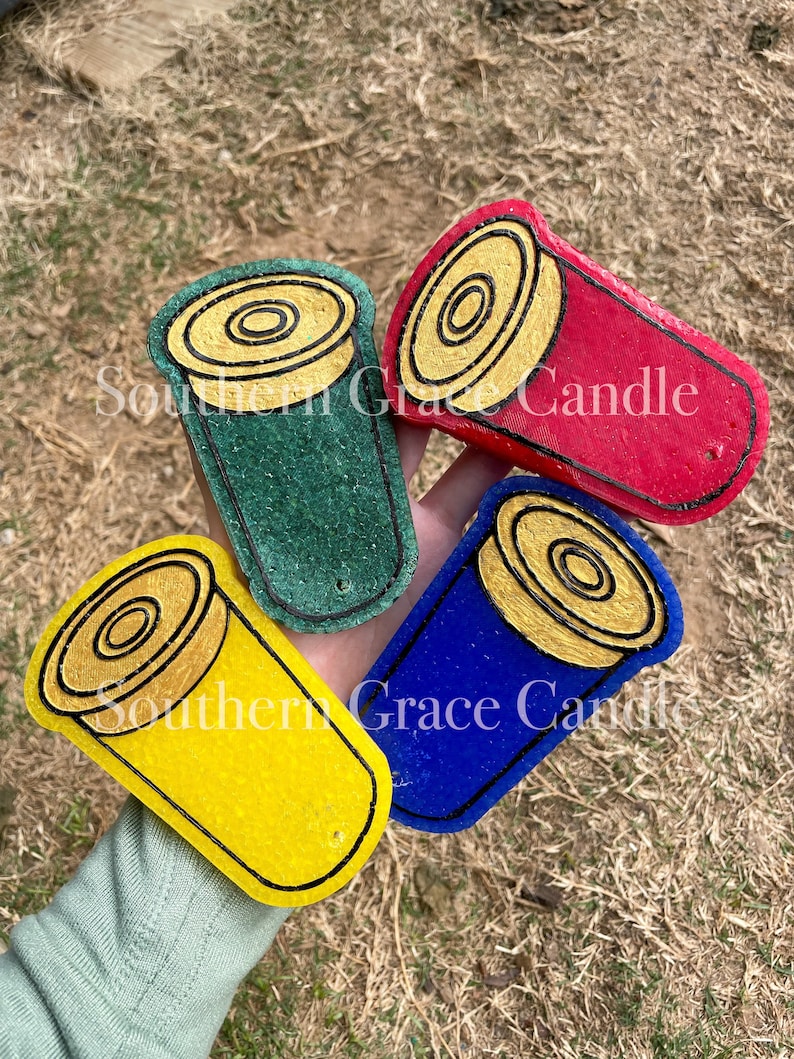 May include: Four shotgun shell candle holders in different colors: green, red, blue, and yellow. Each holder has a gold-colored top with a black outline.
