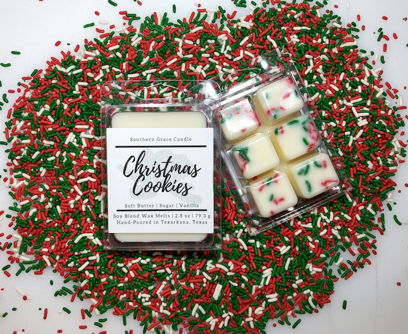 May include: Six white wax melts in a clear plastic container. The wax melts are decorated with red and green sprinkles. The container is surrounded by red, green, and white sprinkles. The label on the container reads "Southern Grace Candle Christmas Cookies Soft Butter | Sugar | Vanilla Soy Blend Wax Melts 2.8 oz | 79.3 g Hand-Poured in Texarkana, Texas".