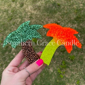 May include: Two palm tree shaped car air fresheners. One is green and brown, the other is yellow, orange, and red.  The text "Southern Grace Candle" is visible in the image.