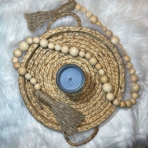 May include: A blue candle sits in the center of a woven tray with a natural rope trim. The tray is surrounded by a garland of wooden beads with natural jute tassels.