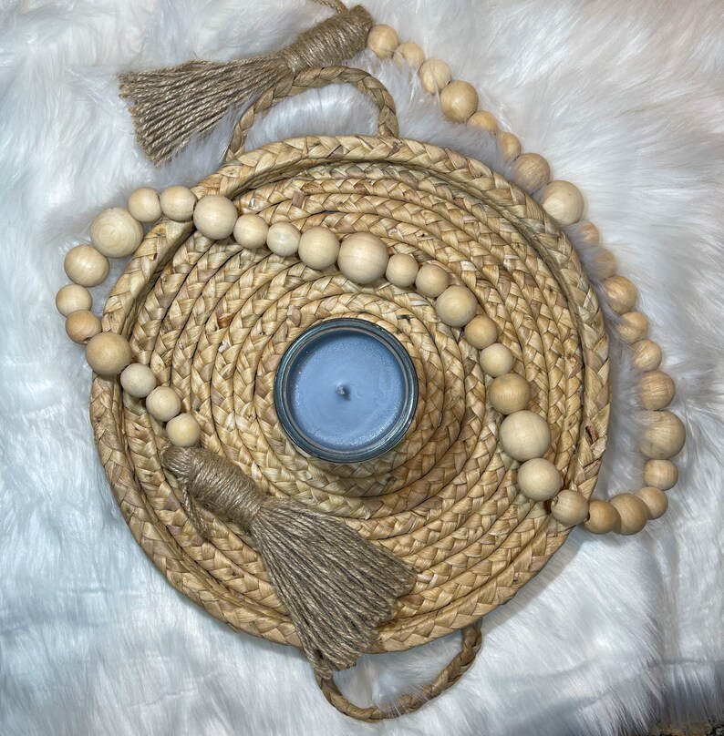 May include: A blue candle sits in the center of a woven tray with a natural rope trim. The tray is surrounded by a garland of wooden beads with natural jute tassels.
